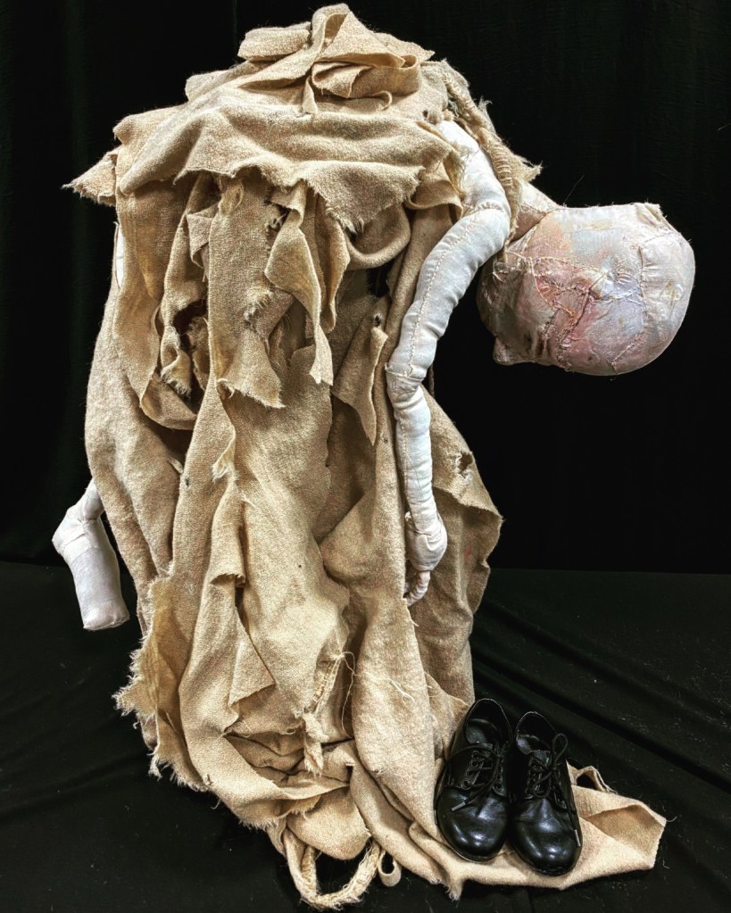 Engulfment ©Teresa Wilson, a soft sculpture doll dressed in rags bending down to pick up a pair of black Oxford shoes.