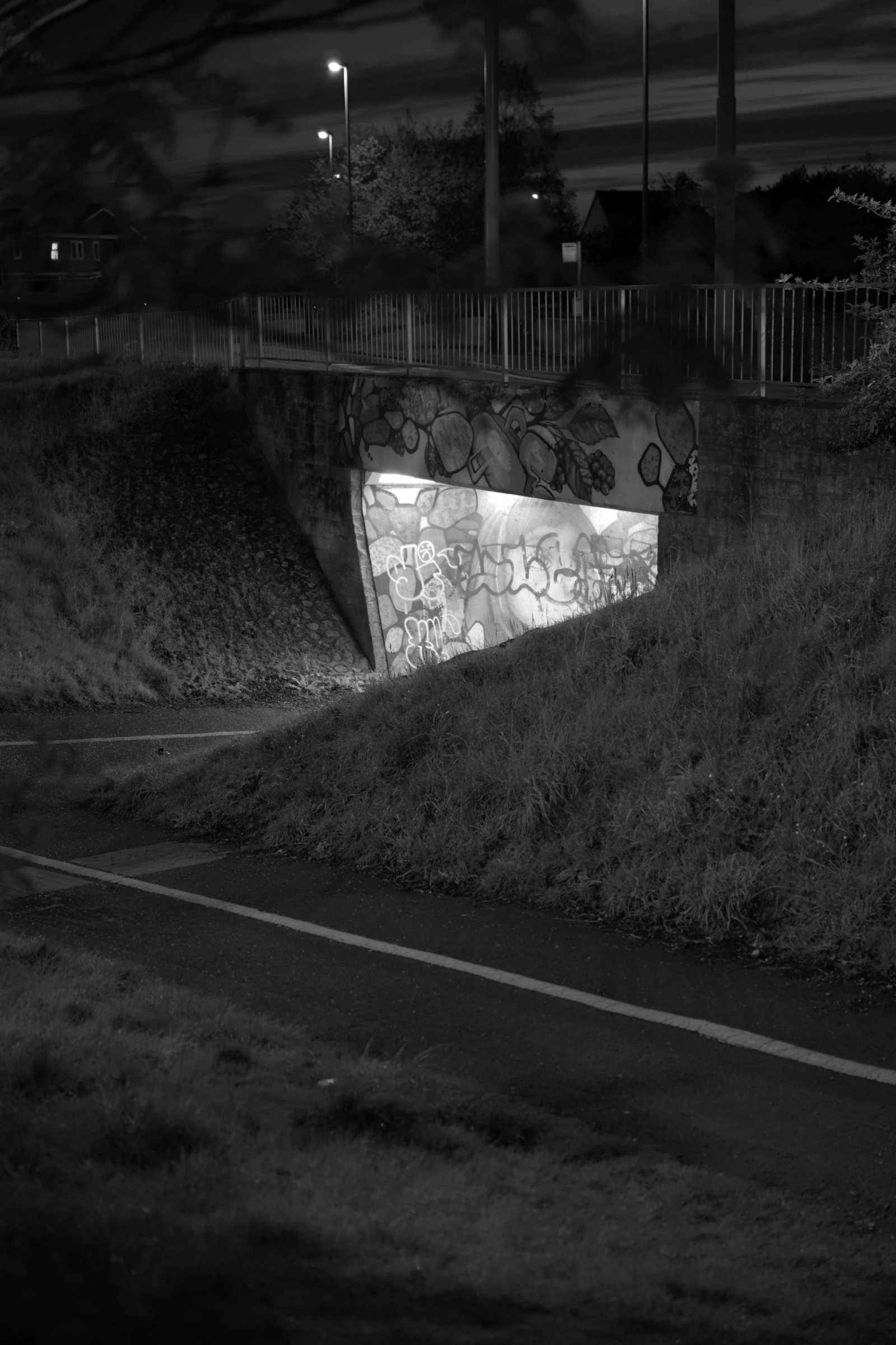 From the series ‘Estate of Magick’ ©Peter Bonnell, a black and white photograph taken at night in a British council estate of a lighted pedestrian underpass with graffiti.